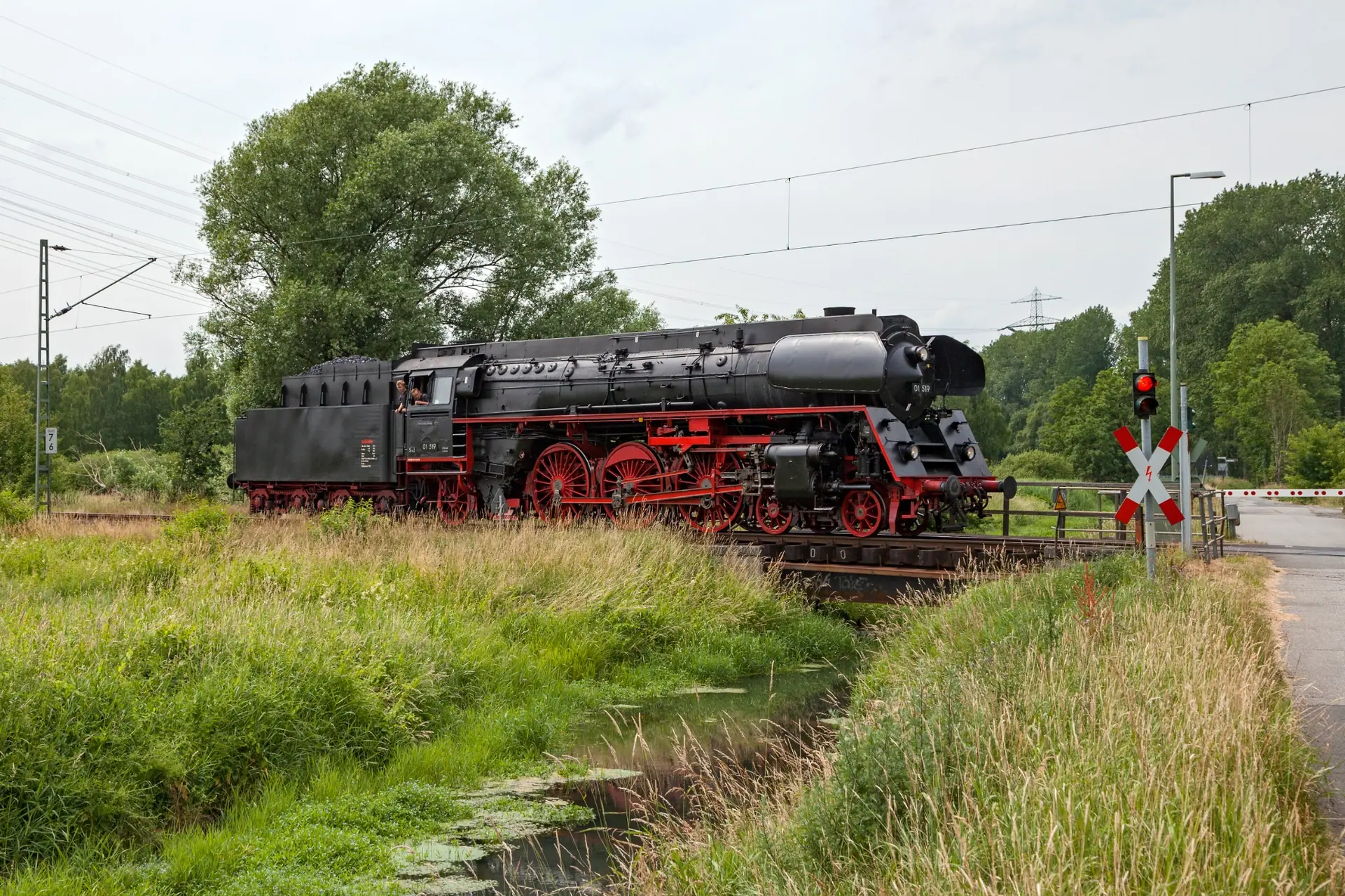 Vorbildaufnahme einer Dampflokomotive an einem Bahn&uuml;bergang mit Graben und hoher Vegetation